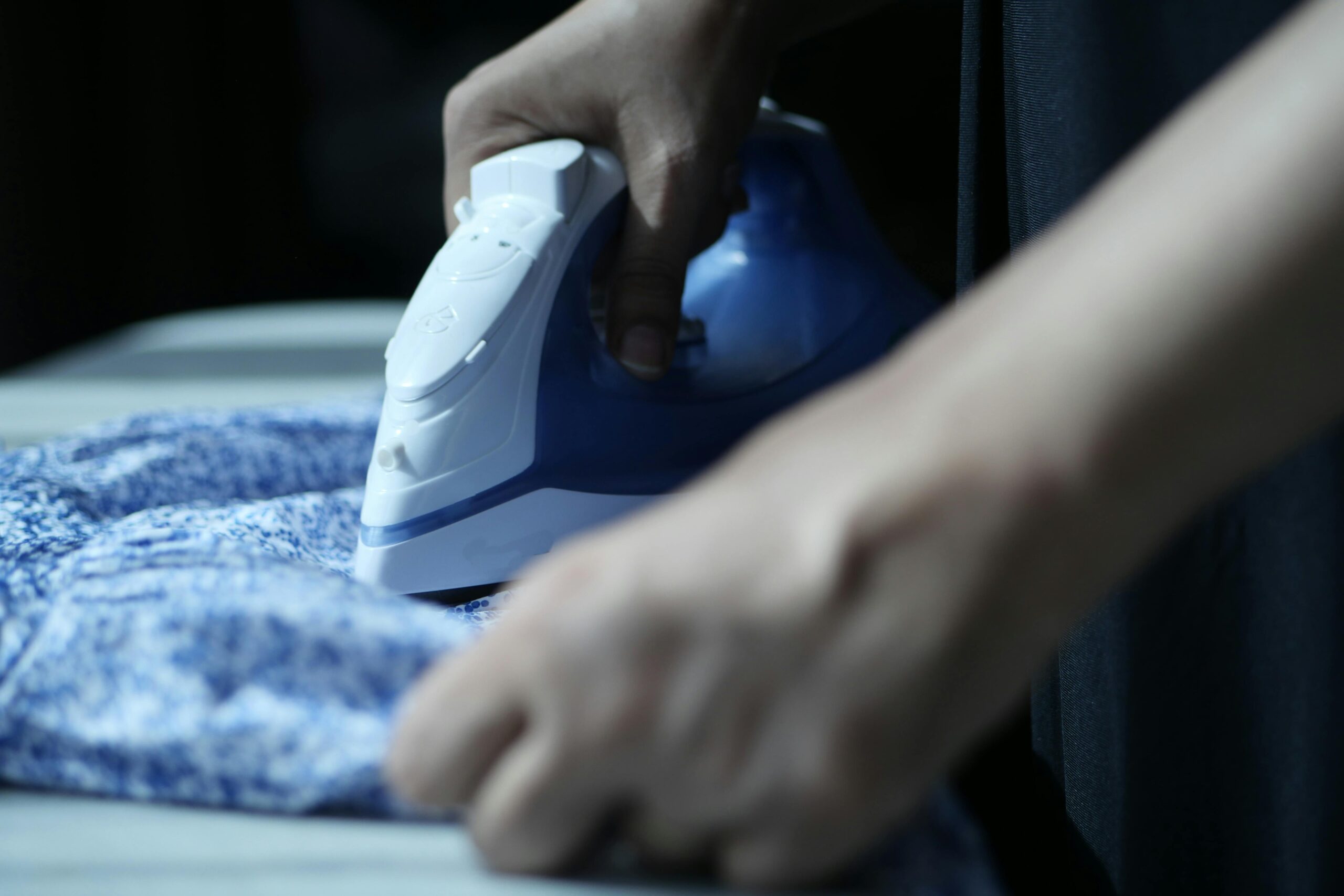 Detailed view of hands using an electric iron on blue fabric indoors.